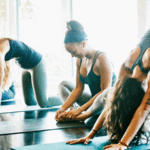 Women practice yoga in a sun-filled room. They are all wearing dark sleeveless tops. From left to right, one is sitting, the next is doing Cat Pose, the third is smiling seated in easy pose, the fourth with a tattoo on her left am is doing Downward Facing Dog