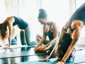 Women practice yoga in a sun-filled room. They are all wearing dark sleeveless tops. From left to right, one is sitting, the next is doing Cat Pose, the third is smiling seated in easy pose, the fourth with a tattoo on her left am is doing Downward Facing Dog