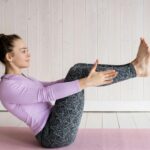 Woman in pink shirt and gray leggings practicing Boat Pose (Navasana) on a pink yoga mat.