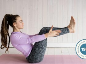 Woman in pink shirt and gray leggings practicing Boat Pose (Navasana) on a pink yoga mat.