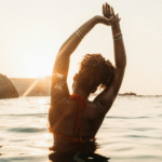 Woman with curly hair pulled back with a tie while she's in the sea and stretching her arms upward