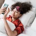 Woman lying in bed with sleep mask on looking at morning meditations on her phone.