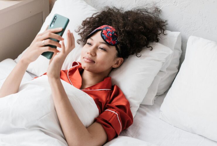 Woman lying in bed with sleep mask on looking at morning meditations on her phone.