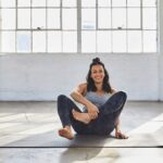 Woman sitting and laughing on a yoga mat as she tries to come into Lotus Pose