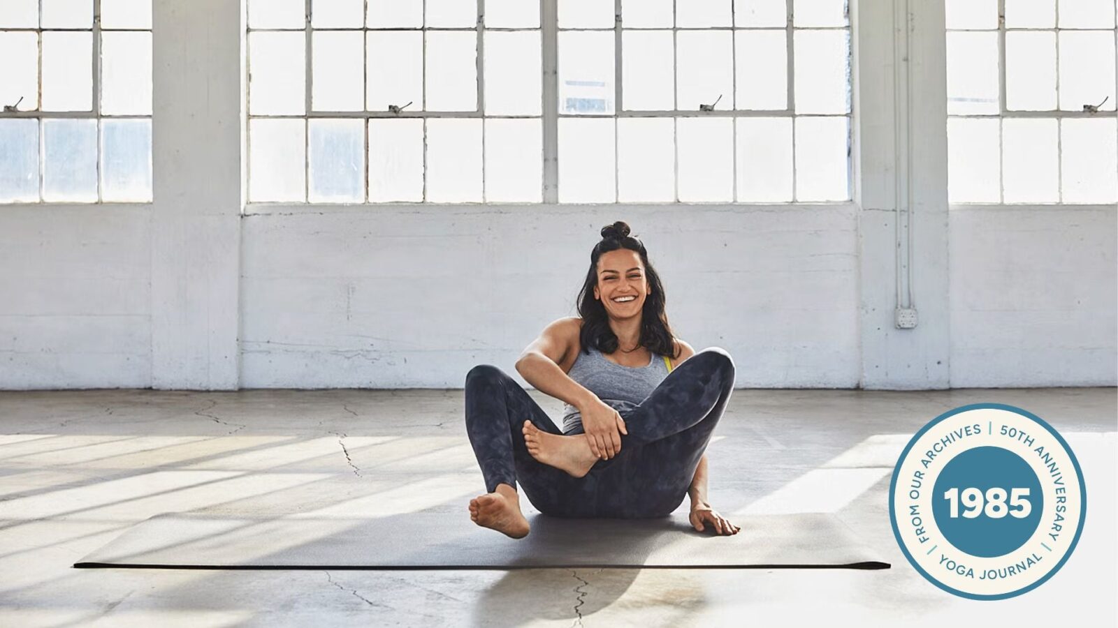 Woman sitting and laughing on a yoga mat as she tries to come into Lotus Pose