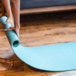 Woman rolling out a blue yoga mat onto a wooden floor.