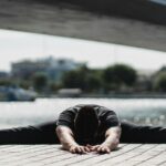 Image of man practicing Wide Legged Seated Forward Fold with a bridge and water in the background.