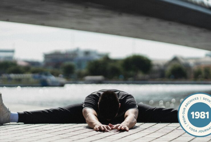 Image of man practicing Wide Legged Seated Forward Fold with a bridge and water in the background.