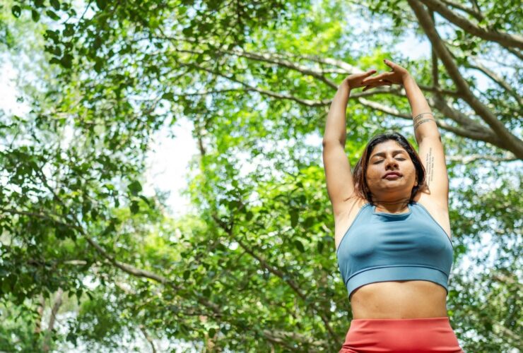 Image of woman in blue sports top and red yoga pants standing outside near green trees reaching her arms up above her head.