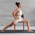 Woman practicing Warrior 2 on a chair against a black and white background of a yoga studio.