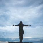 Woman silhouette standing on a cliff near a beach with dark clouds considering the weekly horoscope for November 17-23, 2024.