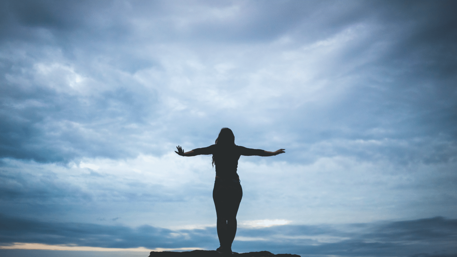 Woman silhouette standing on a cliff near a beach with dark clouds considering the weekly horoscope for November 17-23, 2024.